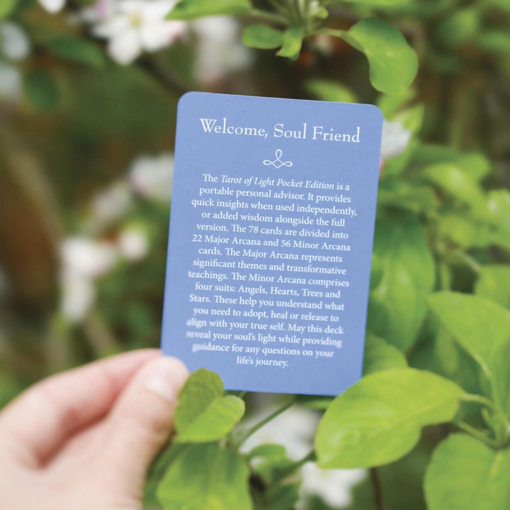A hand holds a blue card titled "Welcome, Soul Friend" in front of green leaves and white flowers. The card appears to be the product being shown, photographed outdoors with a soft blurred natural background.
