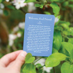 A hand holds a blue card titled "Welcome, Soul Friend" in front of green leaves and white flowers. The card appears to be the product being shown, photographed outdoors with a soft blurred natural background.