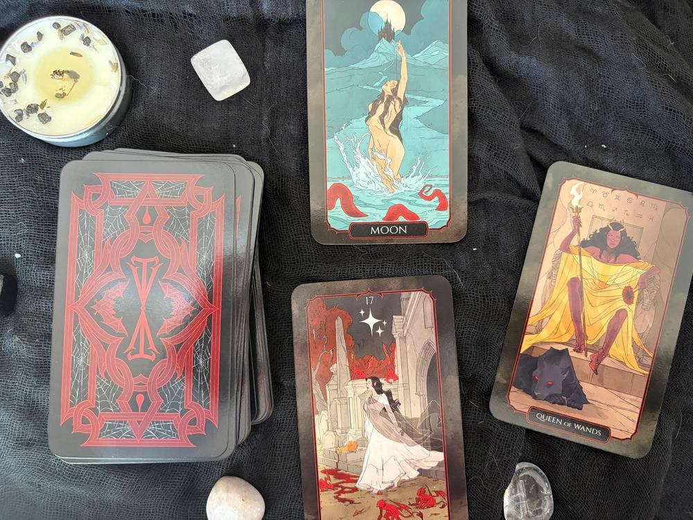 A tarot card deck arranged on a black fabric surface with several illustrated cards visible, including Moon and Queen of Wands, alongside crystals and a candle.