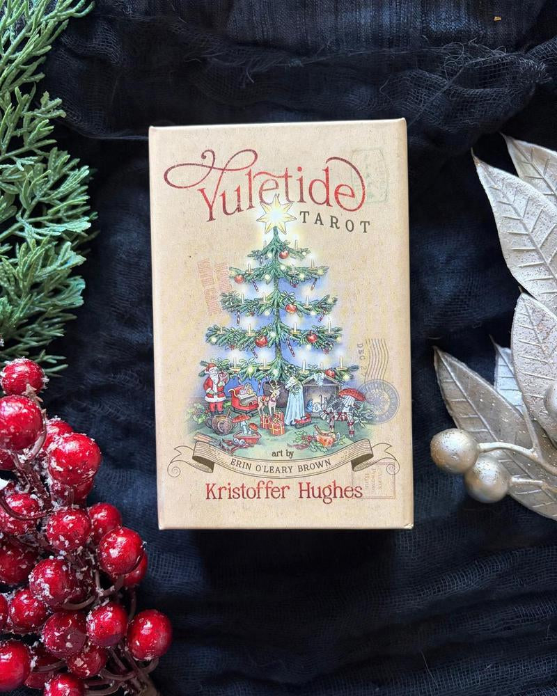 A boxed tarot card deck titled 'Yuletide Tarot' by Kristoffer Hughes, shown standing upright on black fabric with holiday decorations around it, including red berries, green pine branches, and silver leaves.