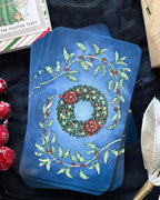 A stack of blue tarot cards featuring a festive Christmas wreath design with holly leaves and red berries, styled on a dark textured fabric with holiday props.