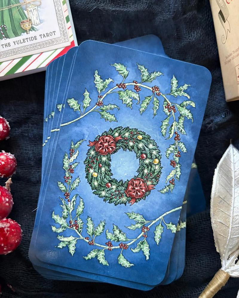 A stack of blue tarot cards featuring a festive Christmas wreath design with holly leaves and red berries, styled on a dark textured fabric with holiday props.