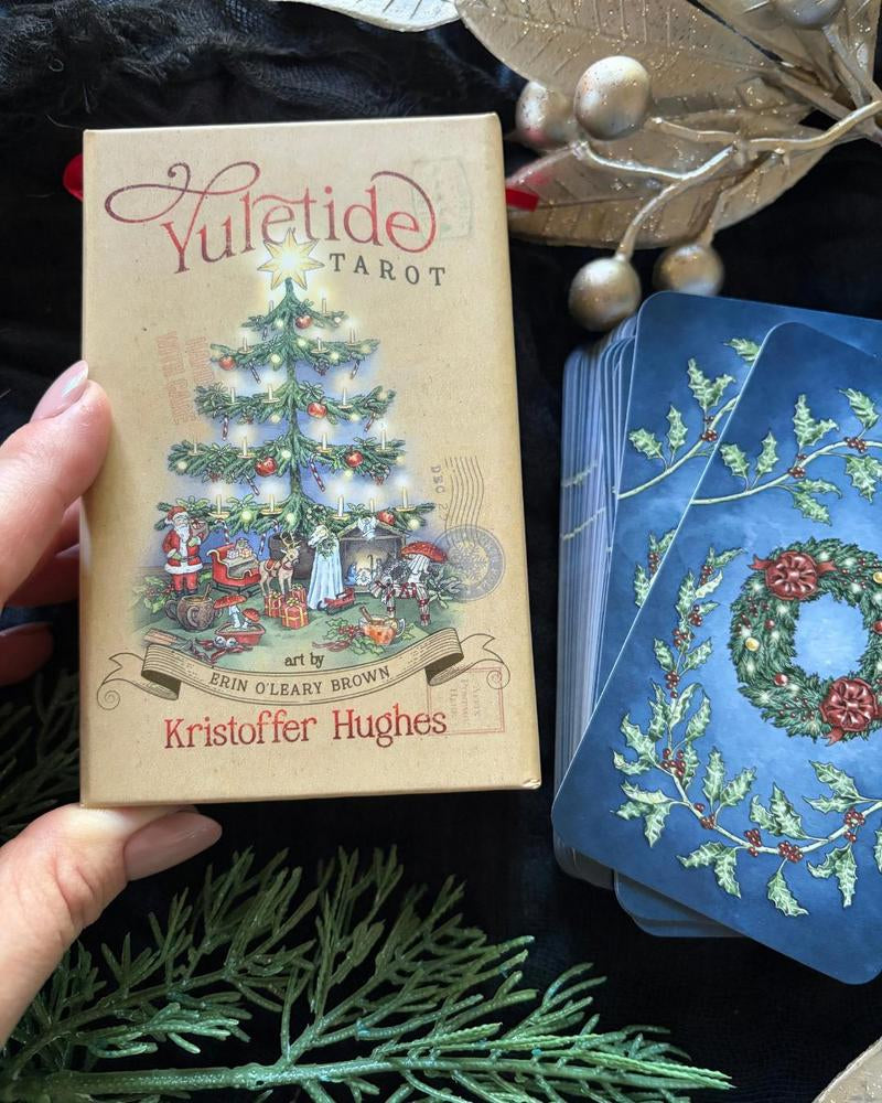 A hand holds a boxed tarot deck titled 'Yuletide Tarot' with a vintage Christmas tree illustration on the box. Blue cards with a holly wreath design are fanned out beside the box on a dark fabric surface, with decorative greenery and metallic ornaments around it.