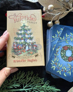 A hand holds a boxed tarot deck titled 'Yuletide Tarot' with a vintage Christmas tree illustration on the box. Blue cards with a holly wreath design are fanned out beside the box on a dark fabric surface, with decorative greenery and metallic ornaments around it.