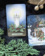 Two illustrated tarot-style cards displayed on a dark fabric background with evergreen branches and gold ornaments. The left card shows a lit candle with holly and the title 'Ace of Candles.