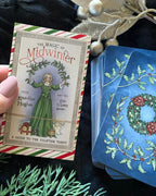 A hand holds the front cover of 'The Magic of Midwinter' Keltide Highher tarot deck, showing vintage-style holiday artwork, next to a fanned stack of blue-backed cards with holly and wreath illustrations, styled on dark fabric with decorative greenery and metallic ornaments.