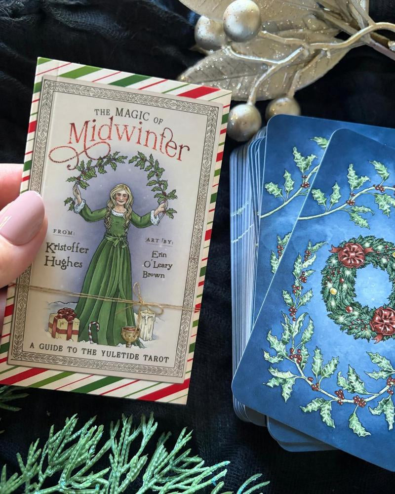 A hand holds the front cover of 'The Magic of Midwinter' Keltide Highher tarot deck, showing vintage-style holiday artwork, next to a fanned stack of blue-backed cards with holly and wreath illustrations, styled on dark fabric with decorative greenery and metallic ornaments.