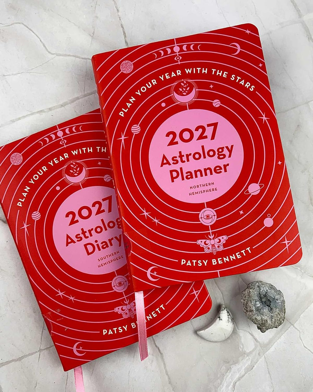 Two red 2027 Astrology Planner notebooks by Patsy Bennett are arranged on a light marble surface with small decorative props, including a white moon and a gray geode.
