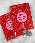 Two red 2027 Astrology Planner notebooks by Patsy Bennett are arranged on a light marble surface with small decorative props, including a white moon and a gray geode.
