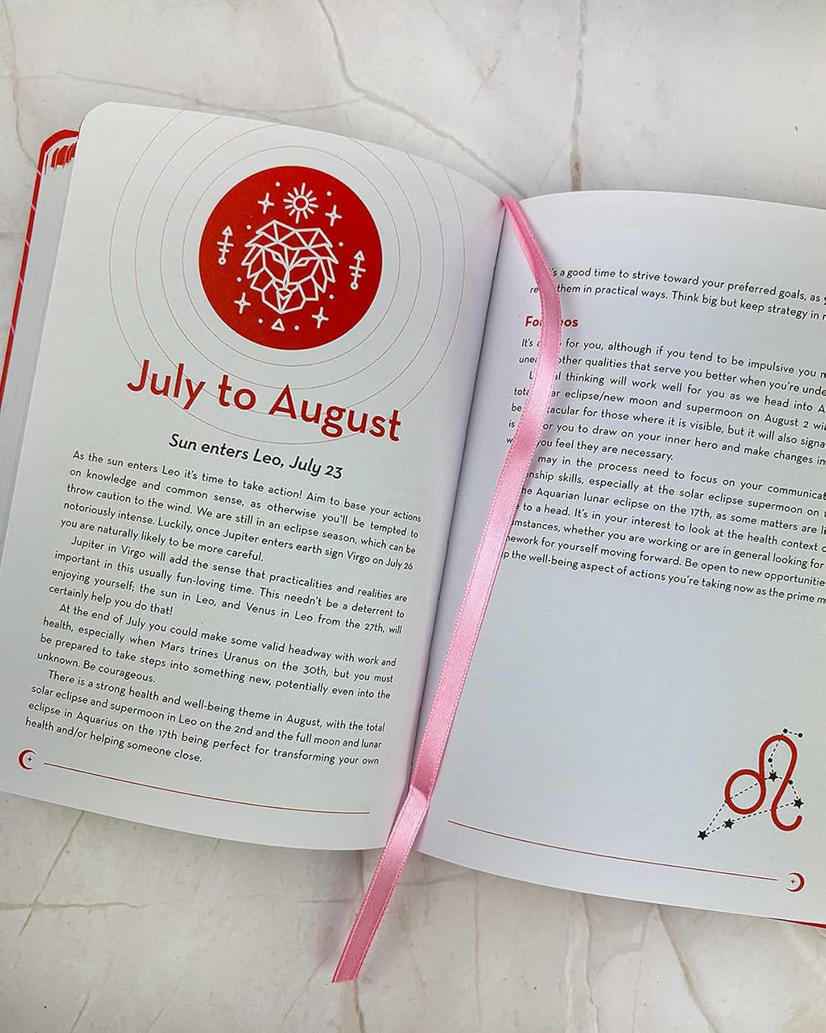 An open book with printed text and red decorative illustrations, featuring a pink pen placed across the pages, photographed on a white marble-like surface.