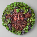 Decorative wall piece with a face surrounded by green leaves and purple grapes on a gray background