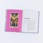 An open book or magazine laid flat on a white background, showing a bright pink left page with a tarot-style 'The Devil' card and a white right page with text titled '15 THE DEVIL'.